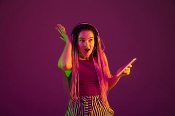 Shocked with headphones, tablet. Young caucasian woman on pink studio background in neon light. Beautiful model with dreadlocks. Human emotions, facial expression, sales, ad concept. Freak's culture.