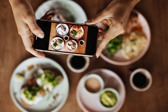 Snapshot of tanned female hands holding smartphone and taking photos of plate with meal. Picture of delicious colorful food on wooden table