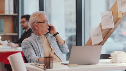 Senior male architect in glasses and casual outfit sitting at workplace in comfortable open space office, typing on laptop and speaking on mobile phone - Powered by Adobe
