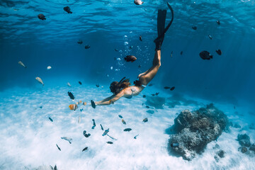 Freediver young woman with fins glides over sandy bottom with tropical fishes in transparent ocean