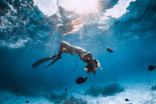 Freediver young woman with fins glides over sandy bottom with tropical fishes in transparent ocean