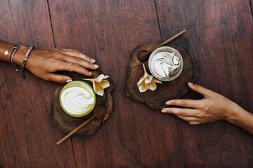 Photo of women hand holding wooden boards with yellow tropical flowers. Snapshot of cup of latte and glass of matcha green tea with milk
