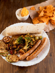 Grilled vegetables and pita with sausage and Mexican corn chips nachos lie on a wooden table. Vertical orientation