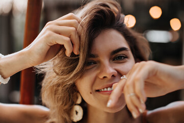 Fototapeta premium Attractive short-haired woman with beautiful tan looks into camera in good mood. Portrait of blonde girl in white handmade earrings posing outside