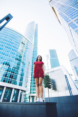 View from below of a young brunette model with perfect figure posing against tall skyscrapers and sky, attractive Caucasian woman with cool style standing in urban setting near modern office buildings