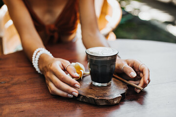 Fototapeta na wymiar Tanned woman in brown bra sits in cafe, poses with glass of coffee with milk. Photo of female hands holding latte and wooden tea spoon