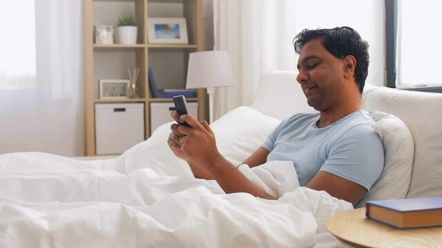 people, bedtime and rest concept - happy smiling indian man with smartphone lying in bed at home