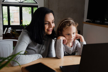 Happy mother and kid daughter looking at laptop, smiling mom and child girl having fun with online video on computer