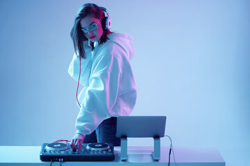 Cool young girl DJ mixes music on a mixing console and laptop, in stylish clothes, glasses on a...