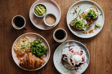 Photo of plates with pancakes,ice cream and strawberry sauce, guacamole, croissant, scramble eggs, sandwich, cups of coffee on wooden table