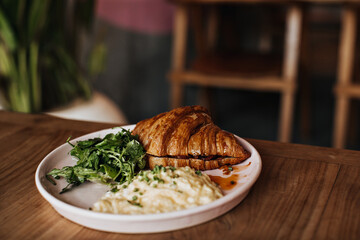 Photo of tasty croissant, scrambled eggs, greens on pink plate on wooden table. Snapshot of tasty food