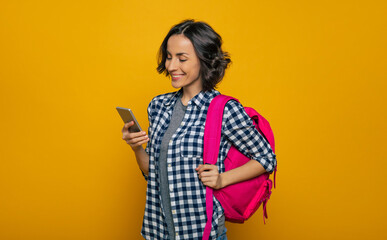  What's new on the internet? A half-length photo of a young beautiful student, dressed casually, with her cute pink backpack on her one shoulder, looking with a wide smile in her smartphone.