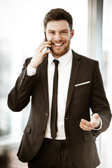 Business concept. Happy smiling young businessman standing in office talking on a cell phone getting good news about his work. Man in suit indoors on glass window background