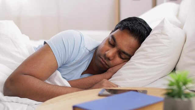 people, bedtime and rest concept - sleepy indian man looking at smartphone in bed at home and goes back to sleep