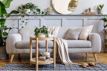 Scandinavian living room interior with design grey sofa, wooden coffee table, tropical plants, shelf, mirror, furniture, plaid pillow, teapot, book and elegant personal accessories in home decor.