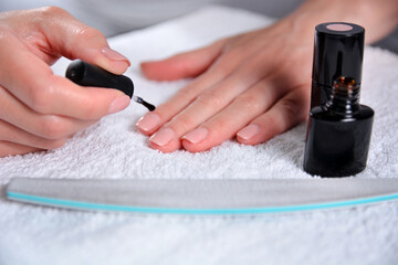 Woman applying nude natural nails polish color at home on a white towel on a desk with a bottle and file. Process of paint nails. Manicure at home and femininity concept. Close up, selective focus