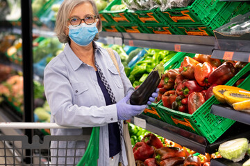 Defocused woman wearing face mask and rubber gloves shopping and choosing fresh vegetables in supermarket, healthy eating, active retired elderly people