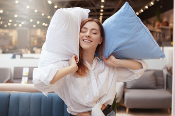 Lovely woman smiling joyfully while shopping for bedding at furniture store. Woman holding pillows,...