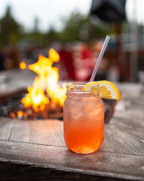 A fruity cocktail drink with a lemon slice served on a mason jar, with a table top fire pit out of focus in the background