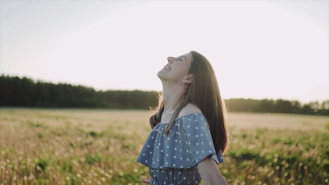 A caucasian beautiful wonan standing on the beautiful field during summer sunset and looking at the camera and then she looking on the sky. The woman wears beautiful dress and. Close up. SLOW MOTION