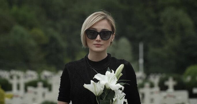Portrait of girl in sunglasses and mourning clothes looking to camera. Young widow holding white lily flower while standing at cemetery. Concept of memorial day. Blurred background