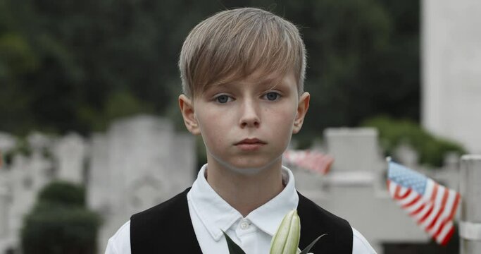 Close up view of teenger kid looking to camera while standing in row of stone crosses with american flags on them. Sad boy at cemetery. Concept of memorial day