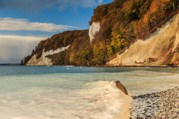 Chalk cliffs on the island of Ruegen. Coast in the morning in autumn at sunrise with sunshine. Trees and stony coast with leaves and clouds in waves of the Baltic Sea in the Jasmund National Park