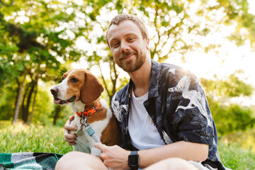 Image of young man smiling and sitting with beagle dog in summer park
