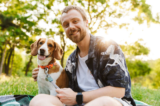 Image of young man smiling and sitting with beagle dog in summer park