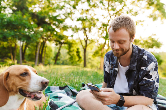 Image of young man using cellphone while sitting with beagle dog in park