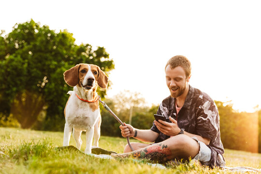 Image of young man using cellphone and sitting with beagle dog in park