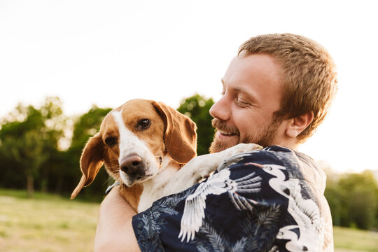 Image of young man smiling and sitting with his beagle dog in park