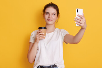 Brunette female with knot holding takeaway coffee and taking selfie via modern smart phone, woman wearing white casual t shirt, standing against yellow wall.