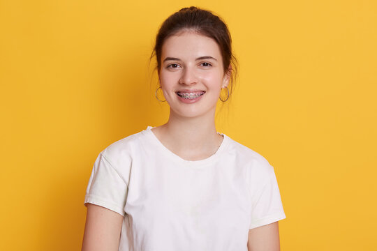 Smiling female with brackets and rounded earrings posing against yellow wall, woman with dark hair looking at camera with happy facial expression, wearing white t shirt.