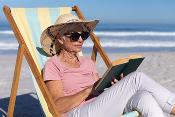 Senior Caucasian woman sitting on a deck chair and reading at the beach.