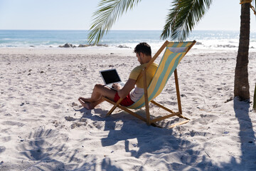 Caucasian man  sitting on a deck chair and using a laptop at the beach