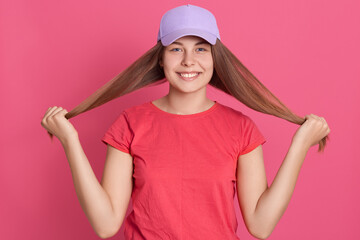 Obraz na płótnie Canvas Happy smiling woman wearing red t shirt and baseball cap pushing her hair aside, posing with toothy smile, standing isolated over pink background.