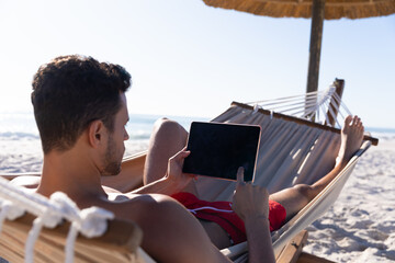 Caucasian man lying on a hammock and using a digital tablet at the beach.