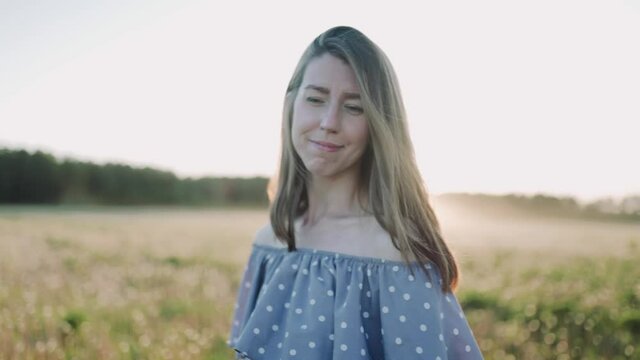 A caucasian beautiful wonan standing on the beautiful field during summer sunset and looking at the camera. The woman is dancing. The woman wears beautiful dress. Close up.