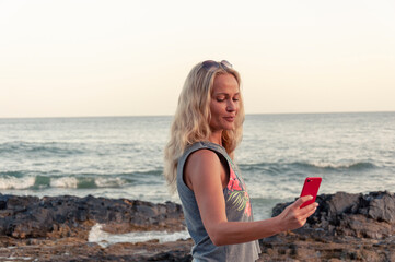 Blonde girl smiles on a rocky seashore and takes a selfie during sunset.