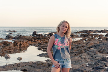 Girl dreamily looks into the lens with her head slightly tilted to the side against the background of the sea and the rocky shore