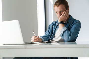 Photo of thinking man writing down notes while working with laptop