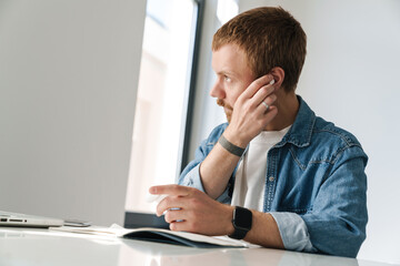 Naklejka premium Photo of ginger man using wireless earphones while working with laptop
