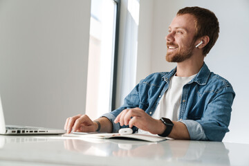 Photo of smiling man using wireless earphones while working with laptop