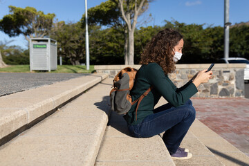 Caucasian woman wearing a protective mask out in the streets, sitting and using her phone