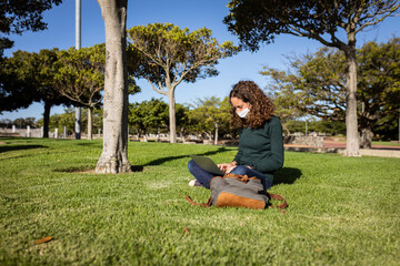 Caucasian woman wearing a protective mask out in a garden and using her laptop