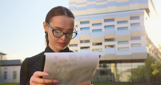 Close-up shooting. The businesswoman is looking through the documents and smiling. She has a folder of documents in her hands. She is standing on the street and in the background is a business center