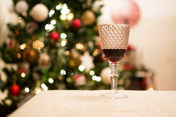 A glass of red wine on the table where the Christmas dinner will be, with the Christmas tree and its lights in the background.