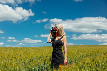 Beautiful blonde girl with suitcase and vintage camera in wheat field in sunny day