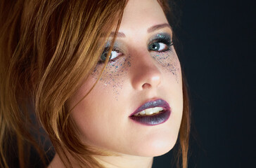 Close up of a young red headed woman with dark make up mascara, purple lip color and metallic colorful glitter under her gray eyes with a charming glance.